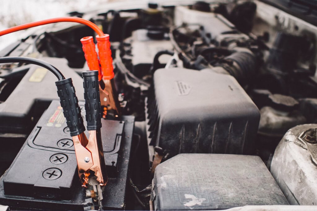 Close-up of a car battery with red and black jumper cables attached, connecting it to another vehicle engine for a jump-start—a crucial service covered by many roadside assistance plans.