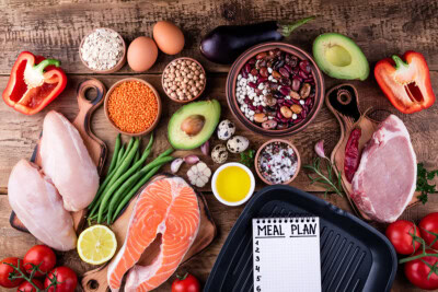 Assorted healthy foods including chicken, salmon, green beans, lentils, chickpeas, eggs, avocado, red peppers, tomatoes, garlic, eggplant are arranged alongside a meal plan notebook on a wooden table for optimal nutrition and counseling support.