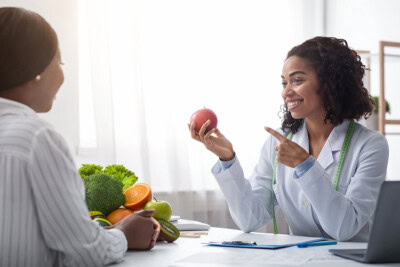 A nutritionist in a white coat holds an apple and engages in nutritional counseling with a patient. Various fruits and vegetables are on the table between them, highlighting the health benefits of a balanced diet.