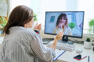 Two women engaged in a video call. One woman is sitting at a desk with a keyboard, clipboard, and smartphone, while the other is seen on the monitor smiling, with plants in the background. Their conversation centers around mental wellness.
