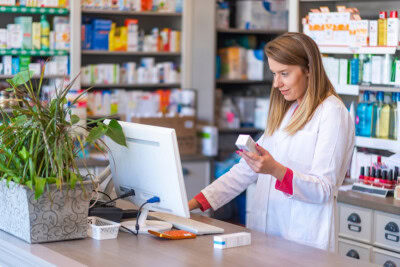 Pharmacist in a white lab coat stands at a pharmacy counter, examining a medication box while using a computer. Shelves stocked with medicines and health products are visible in the background, alongside an Rx Card associated with patient discounts.