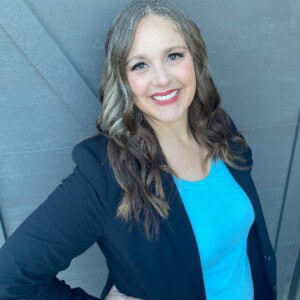 Jessica Fiveash, a woman with long wavy brown hair and a streak of gray, wearing a black blazer over a blue top, smiles at the camera against a gray background.