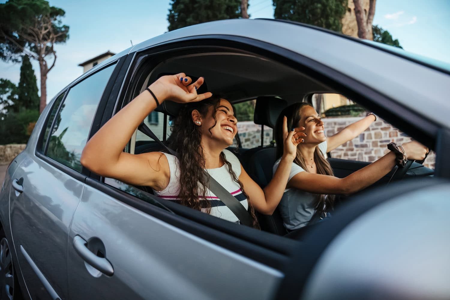 Two young women in a car with windows down, smiling and raising their hands, one is driving and the other in the passenger seat. Trees and a building are visible in the background. Their excitement symbolizes the spirit of entrepreneurship often found in small companies.