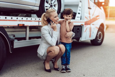 A woman in business attire squats while talking on the phone next to a smiling child standing by a tow truck with a car on it, showcasing the balance families often achieve between work and caregiving.
