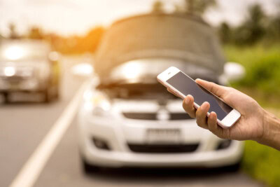 A person holds a smartphone in front of a car with its hood open on the side of the road, possibly waiting for roadside assistance. Another car is visible in the background.