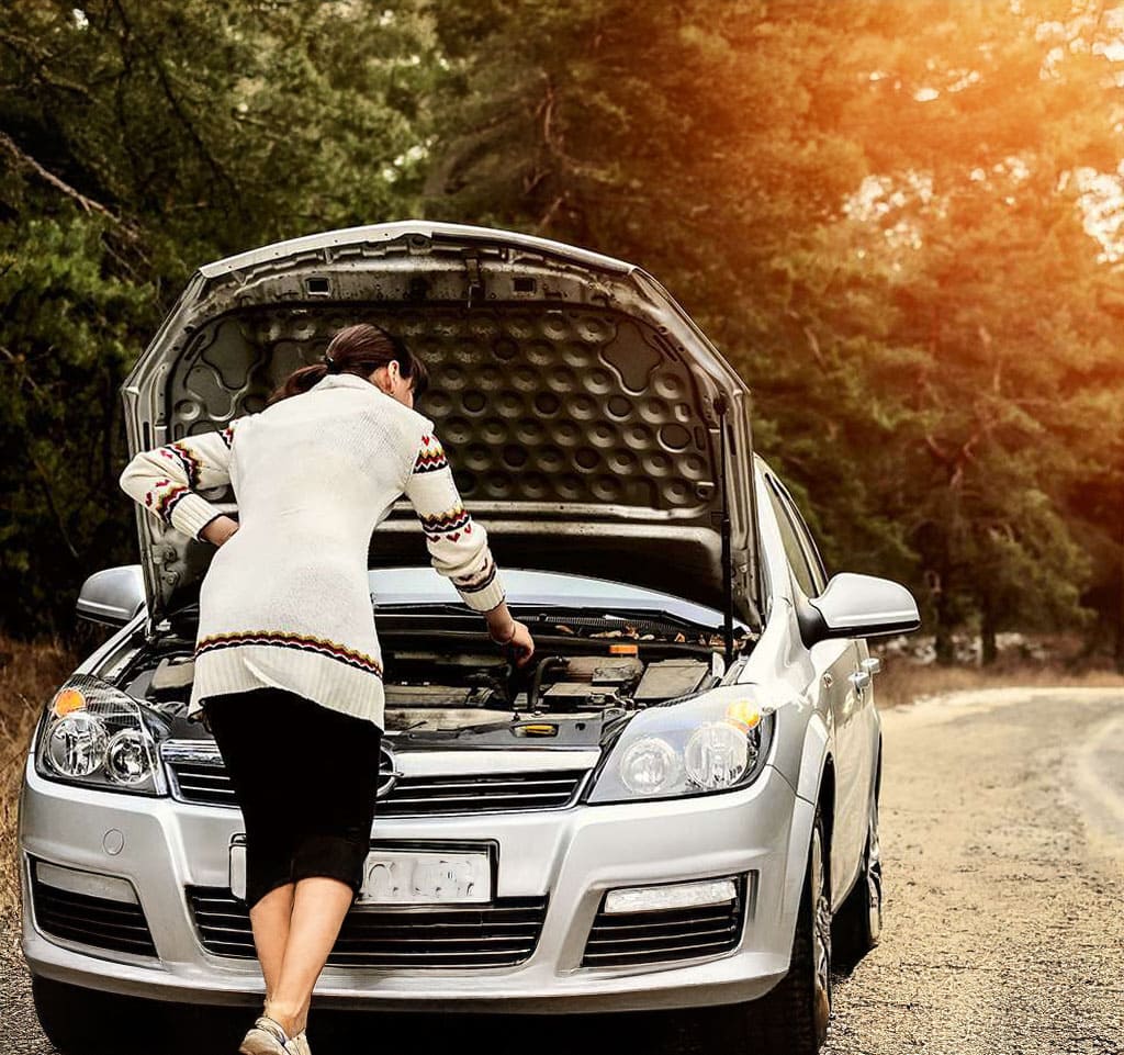 A person wearing a white sweater and black pants checks the engine of a silver car with the hood open on a rural road, likely awaiting Roadside Assistance. Trees are visible in the background with sunlight shining through.