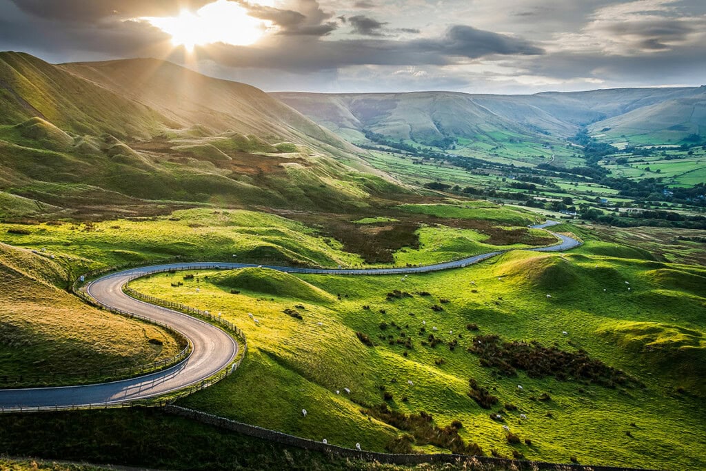 A scenic route winds through lush green hills with a valley in the background, under a sky partly covered by clouds and rays of sunlight breaking through. Perfect for a road trip in the United States.