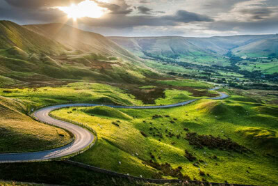 A scenic route winds through lush green hills with a valley in the background, under a sky partly covered by clouds and rays of sunlight breaking through. Perfect for a road trip in the United States.