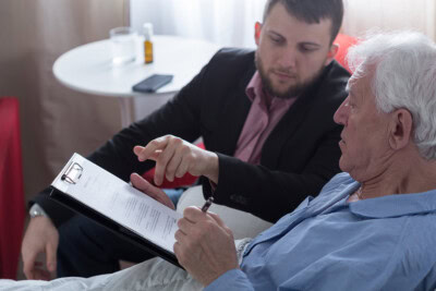 A man offers patient advocacy by assisting an elderly man in filling out paperwork while sitting on a bed. In the background, a table holds a water glass and a bottle.