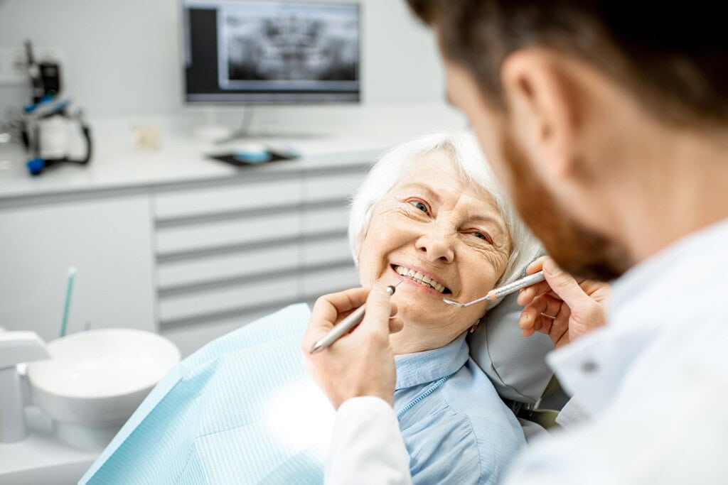 A dentist examines an older woman's teeth using dental instruments as she smiles in the welcoming clinic, which feels almost like home. A computer with dental X-rays is visible in the background.