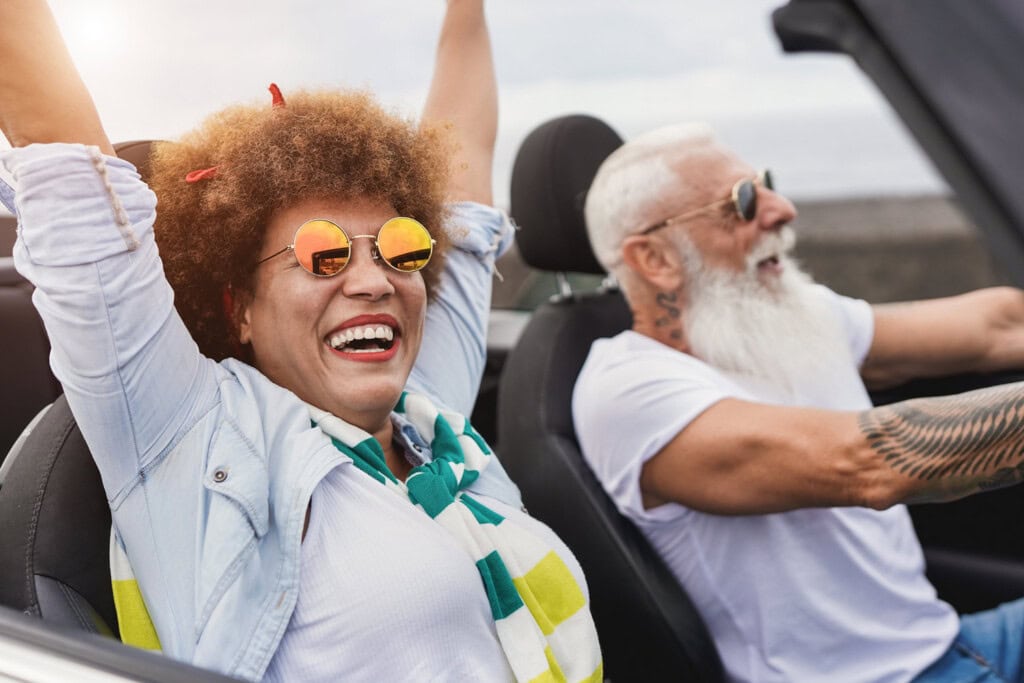 Two seniors ride in a convertible with the top down. The person on the left has arms raised and is smiling, while the person on the right, with a beard and tattoos, is driving and also smiling.