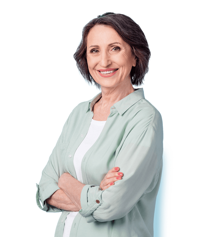 A senior woman with short dark hair, wearing a light green shirt and white top, stands with arms crossed and smiles at the camera.