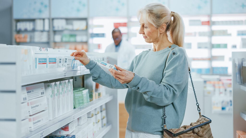 Alliance Health 3 A woman with blonde hair is selecting a product from a shelf in a pharmacy while holding a shopping bag. A person in a white coat is in the background.