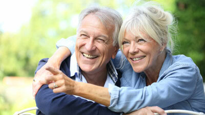 A smiling elderly couple embraces warmly outdoors, with the woman pointing at something in the distance as if to ask, "Who is it for?