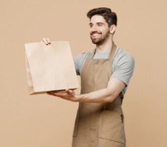 Man with a beard wearing an apron, holding a brown paper bag, and smiling against a beige background, exuding a warm, homey vibe.