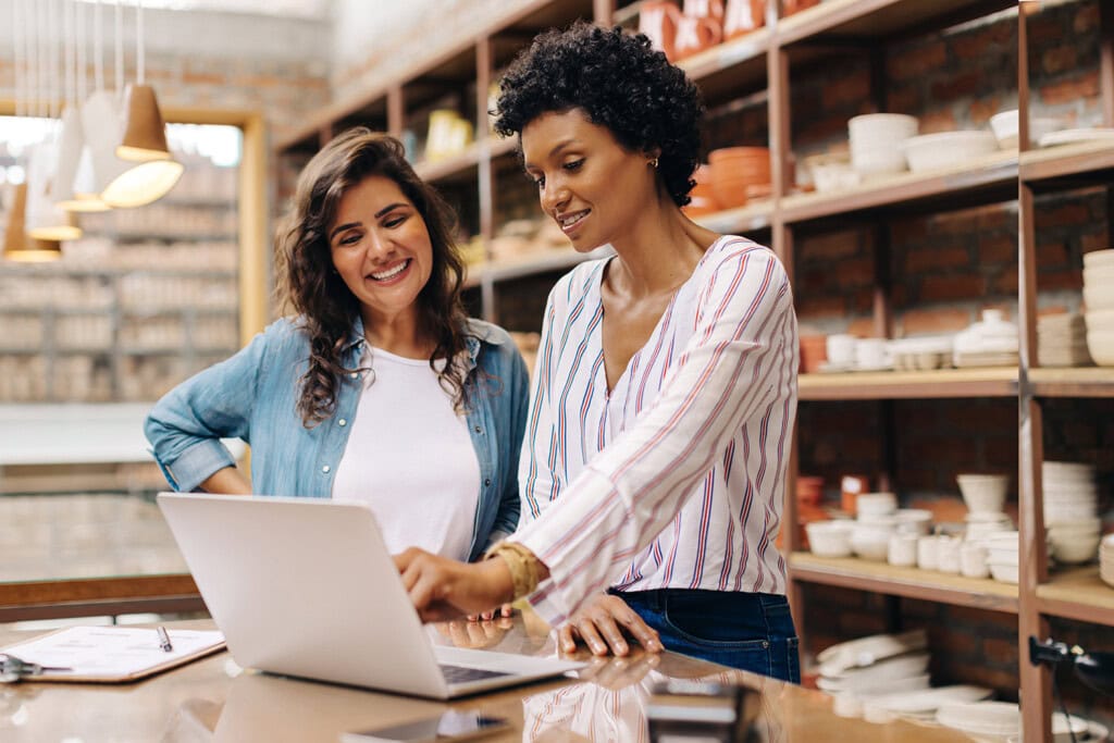 Two women are standing in a workshop, looking at a laptop screen while smiling. Shelves with pottery are in the background, suggesting they might be exploring creative alternatives for small businesses.