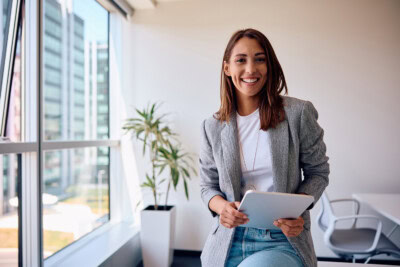 A woman in a blazer holding a tablet stands in a bright office with large windows and a potted plant, reflecting the modern wellness approach embraced by many solopreneurs.