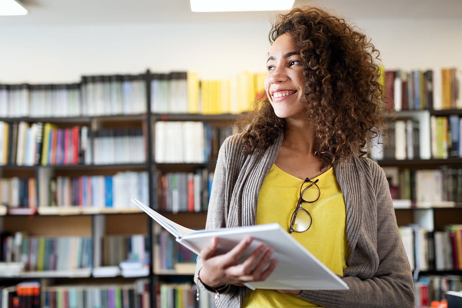 A person stands in a library holding an open book, smiling and wearing a yellow shirt, grey cardigan, and glasses hanging on their shirt. Surrounded by shelves filled with books, they look confident and ready to Get on Track for College.