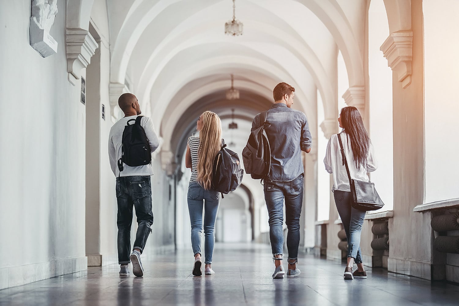 Four College students walk down a university hallway, each carrying backpacks. The arched corridor is well-lit, and they discuss the upcoming $200,000 Question trivia challenge.