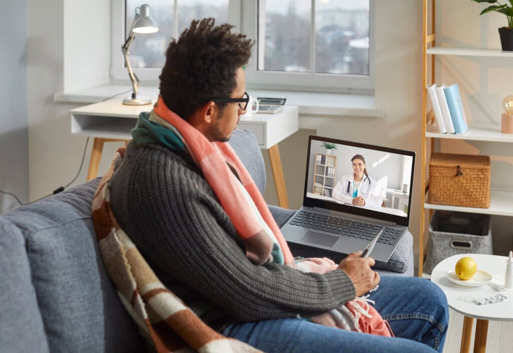 A man sits on a couch wrapped in a blanket, holding a tablet and having a video call with his small business doctor on the laptop placed on the table in front of him.