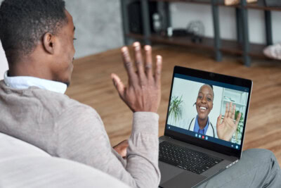 A man sitting on a couch waves at a person on a video call displayed on a laptop screen. Utilizing telemedicine, the person on the screen also waves back.
