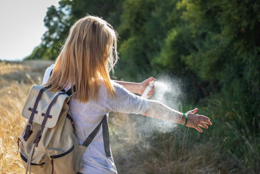 A person with blonde hair and a beige backpack sprays insect repellent on their arm while standing outdoors near trees and tall grass, following safety tips for tick bite prevention.