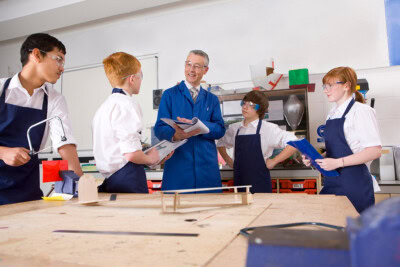 A teacher in a blue lab coat instructs four students wearing safety glasses and aprons in a workshop setting. The students, focused on their career education, hold clipboards and gather around a workbench with wooden models, all funded by a generous grant.
