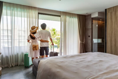 A couple stands by the window in their Alliance Travel hotel room, holding each other with luggage beside them, looking out onto a balcony with a view.