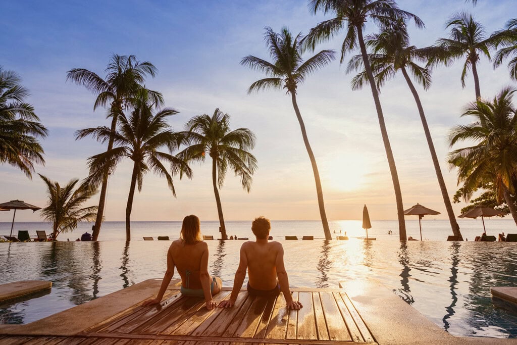 Two people sit by an infinity pool at sunset, gazing at the ocean. Palm trees sway gently, and lounge chairs dot the background. Experience this serene escape with Alliance Travel.